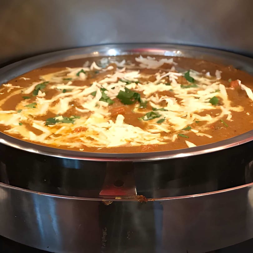 A large metal serving dish filled with a rich, orange curry, topped with white cheese and green herbs.