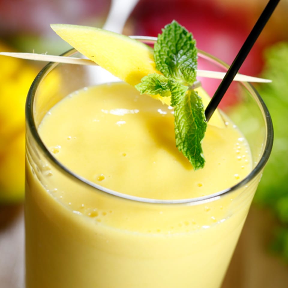 Close-up of a yellow mango smoothie in a glass, garnished with a mango slice and mint.