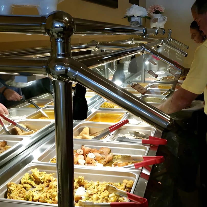 People serving themselves from a buffet line filled with various hot dishes and tongs.